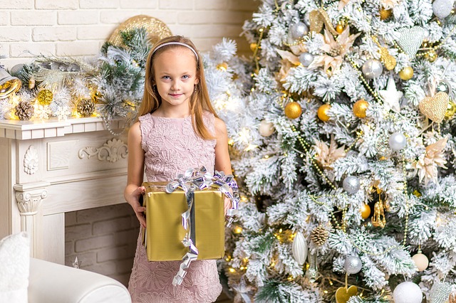 A young girl in a white dress holds a gold wrapped Christmas present in front of a beautiful Christmas tree.